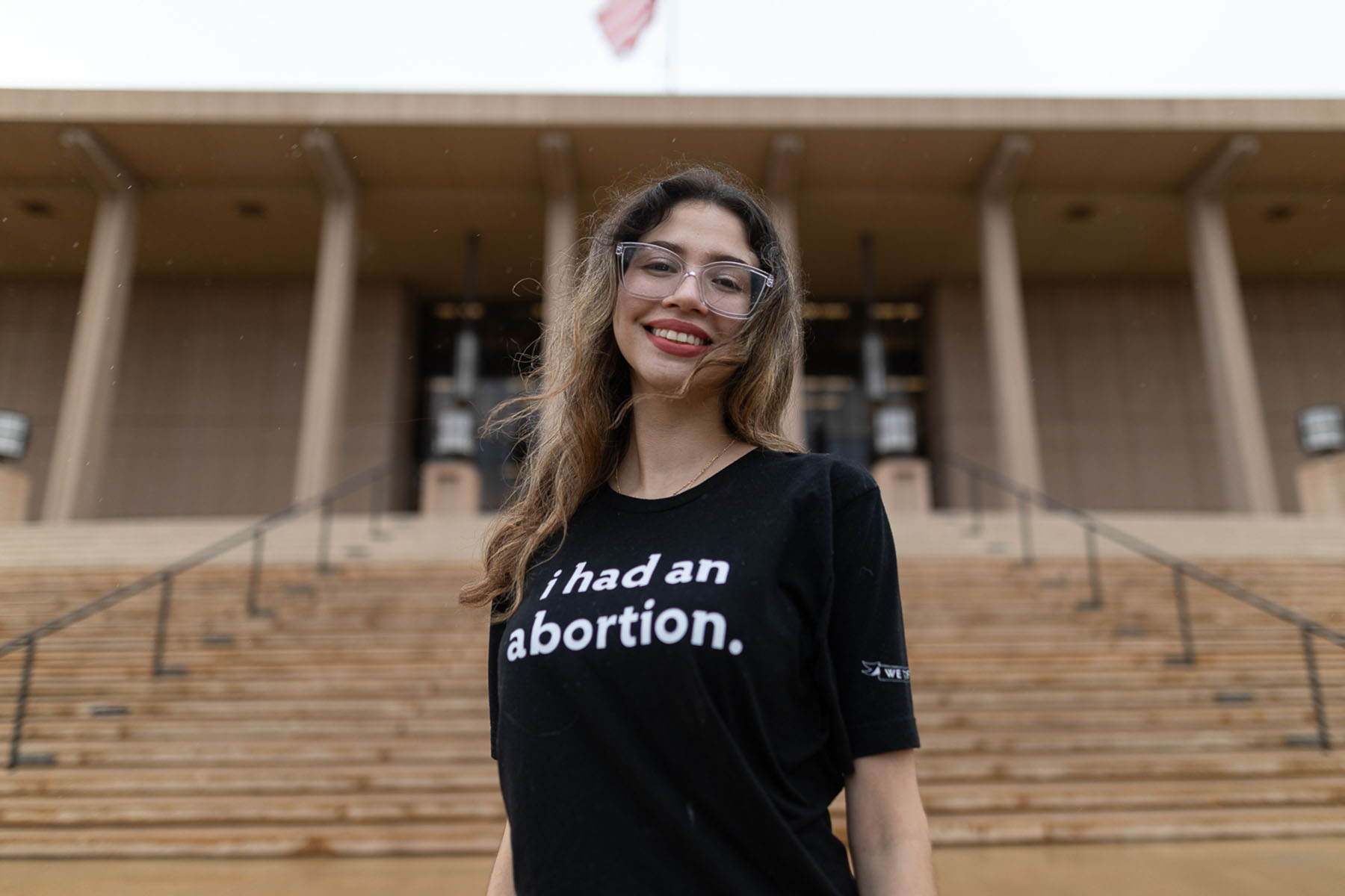 A person with long ashy blonde hair, glasses, wearing burnt orange lipstick and a black shirt that reads "I had an abortion," stands in front of a large building with a staircase.