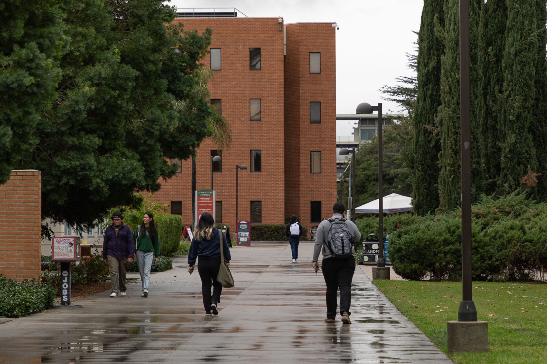 Students walk atop a wet concrete passage way on a campus with brick buildings.