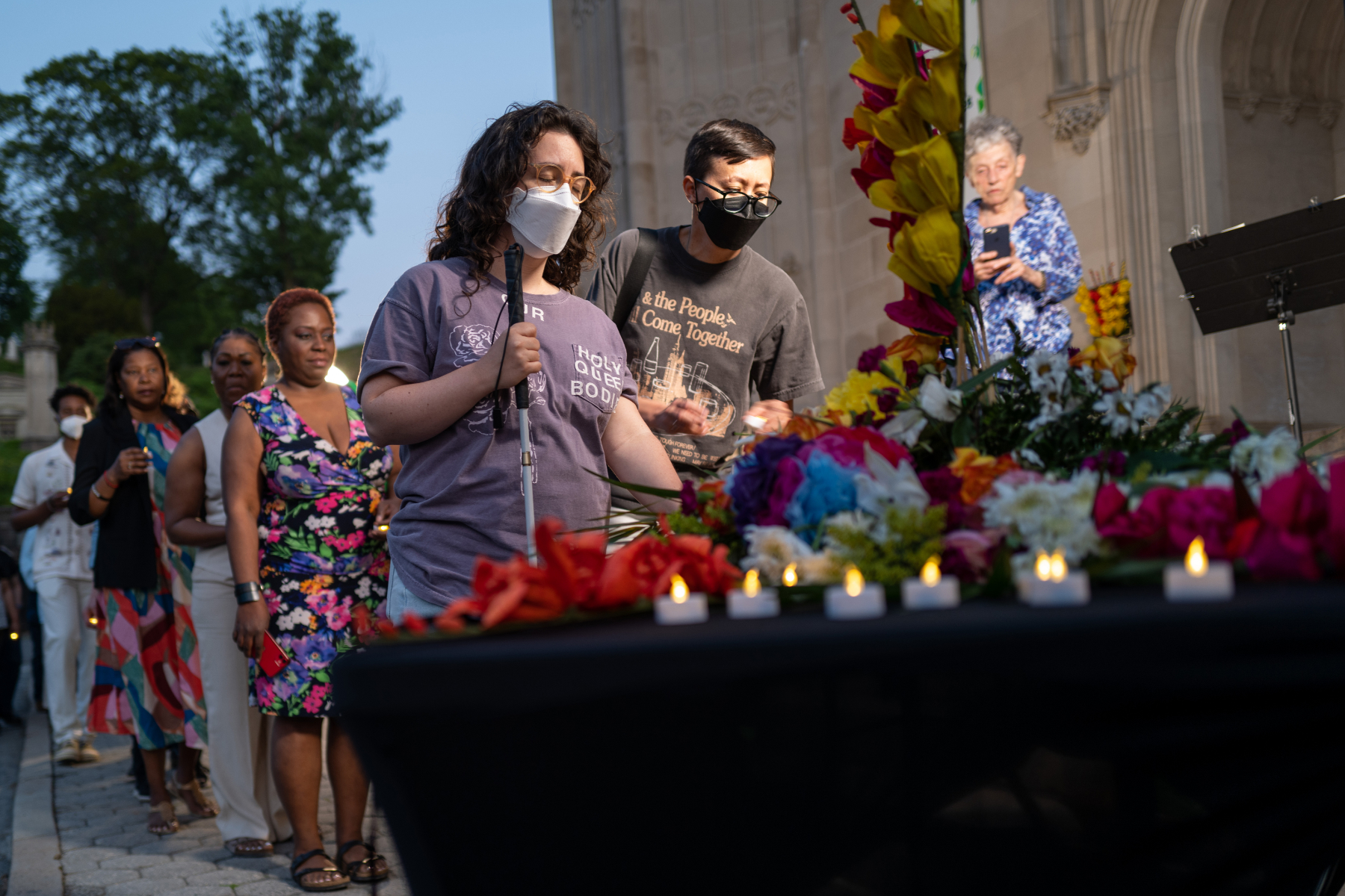 A woman holding a cane and a man in glasses, both wearing masks, are at the head of a line to put candles on a flower-covered memorial table in New York.