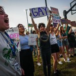 Young LGBTQ+ rights supporters hold signs and protest against Florida Governor Ron DeSantis outside a Trump campaign event.