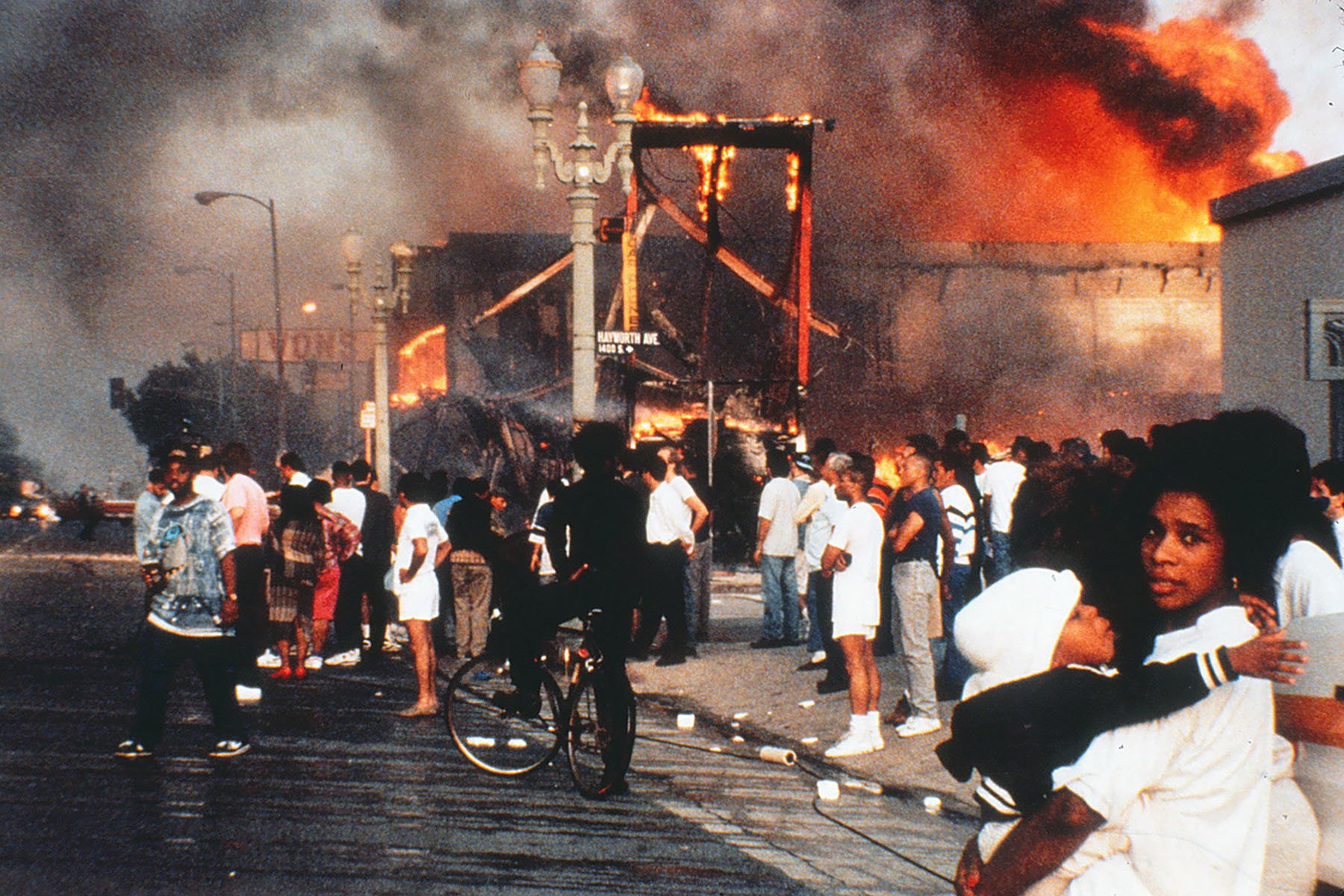 Bystanders watch as a fire rages at an intersection. In the forefront, a mother holds her baby.