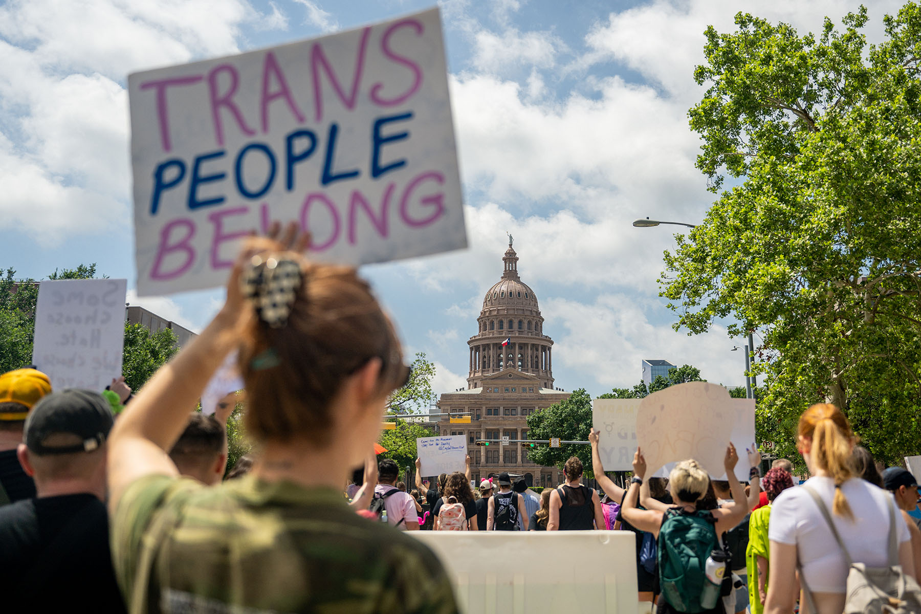 A protesters holds up a sign that reads "Trans People Belong" as the Texas State Capitol can be seen in the background.