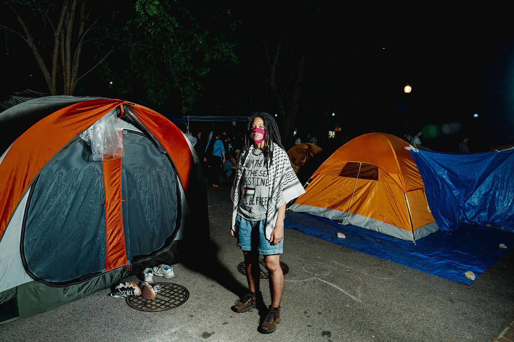 Bobbi-Angelica Morris poses for a portrait amidst tents at the George Washington University encampment. They are wearing a clear face mask, a keffiyeh is draped on their shoulders, and they are wearing a T-shirt with a Palestinian flag on it.