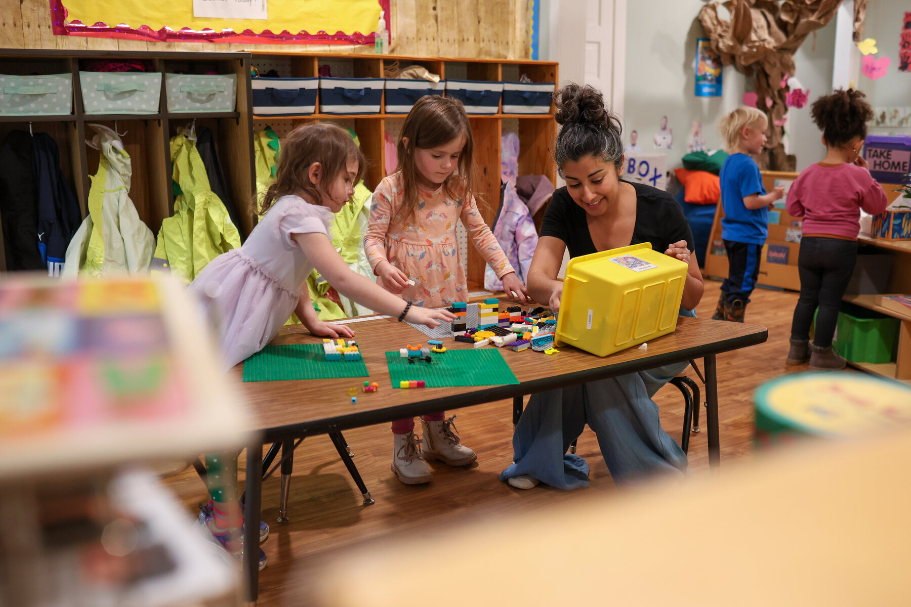 A woman works with two preschool students on a Lego project.