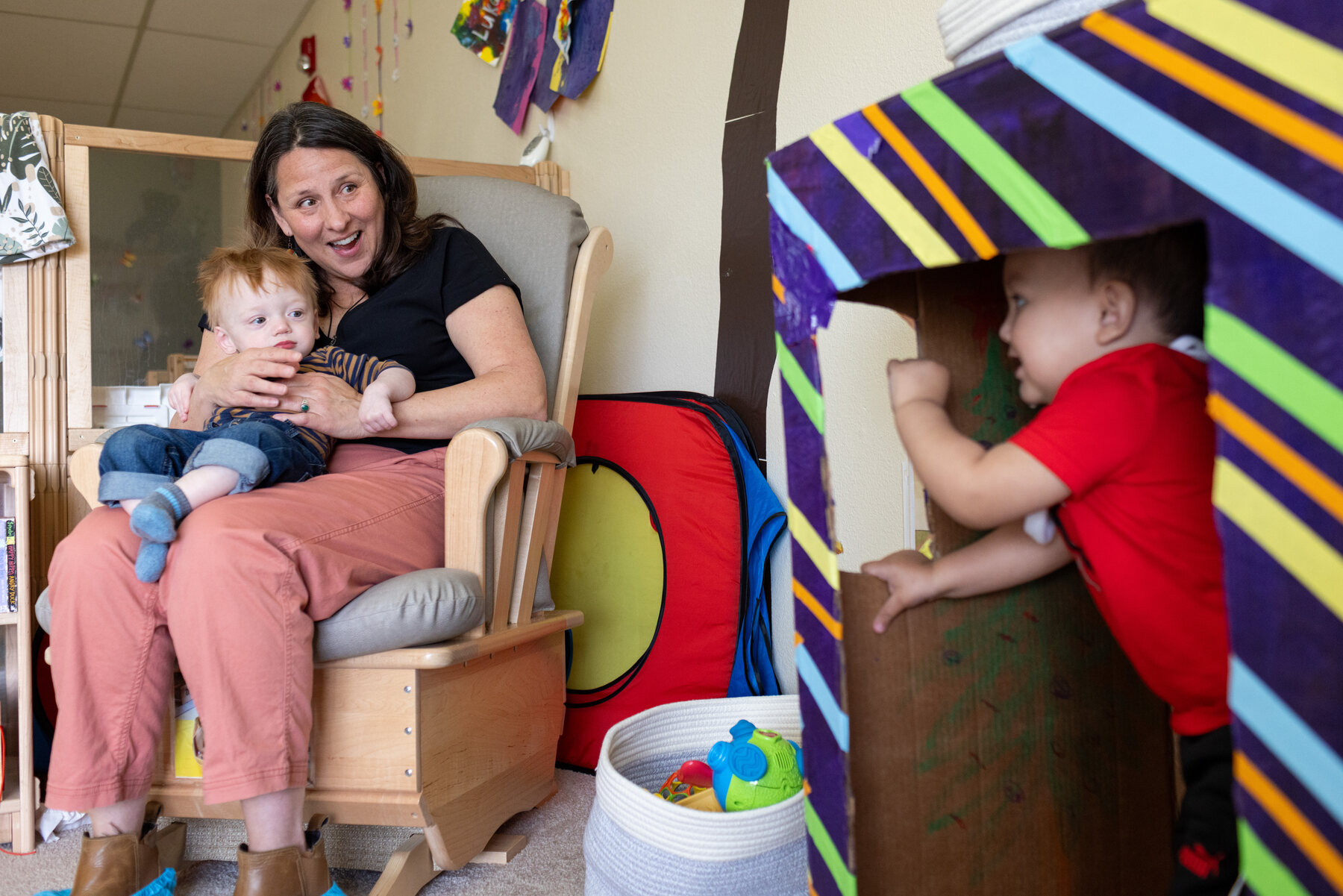 A woman sits in a chair with an infant while a toddler plays next to them.
