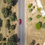 Aerial drone view of a road with vegetation on the sides and a red car driving through it.