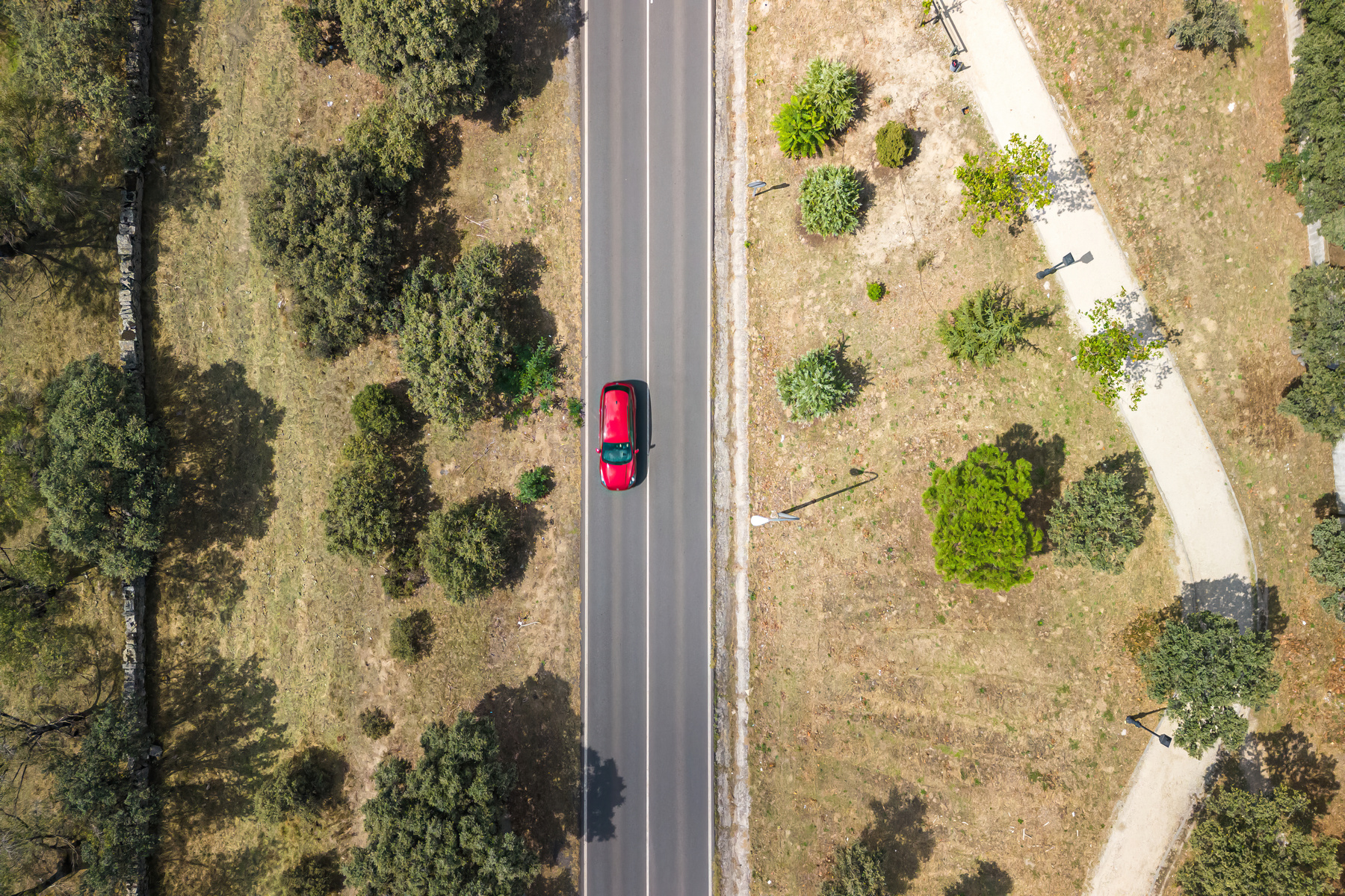 Aerial drone view of a road with vegetation on the sides and a red car driving through it.
