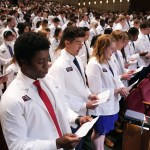 Medical students wearing white coats are seen reading from a paper as they recite an oath in an auditorium.