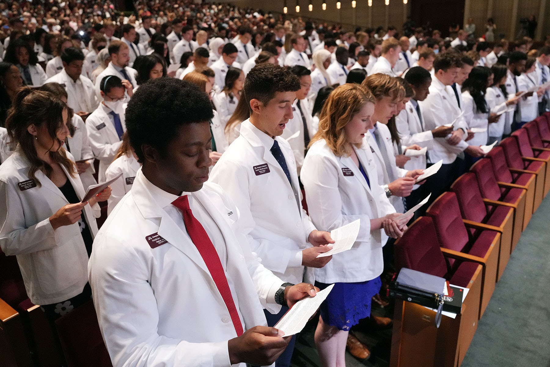 Medical students wearing white coats are seen reading from a paper as they recite an oath in an auditorium.