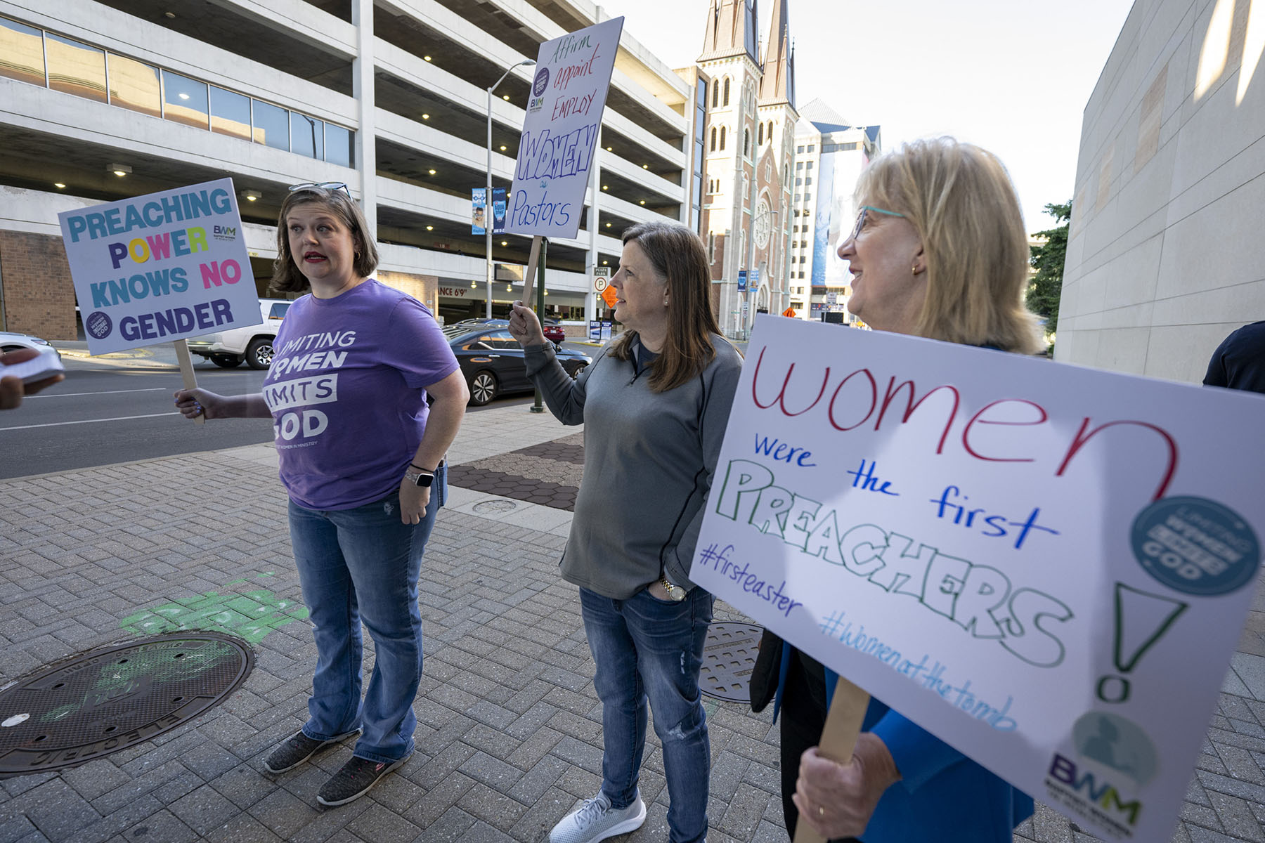 Three women holding signs that read "Preaching power knows no gender," "Affirm, appoint employ women pastors" and "Women were the first preachers" stand outside the Southern Baptist Convention's annual meeting.