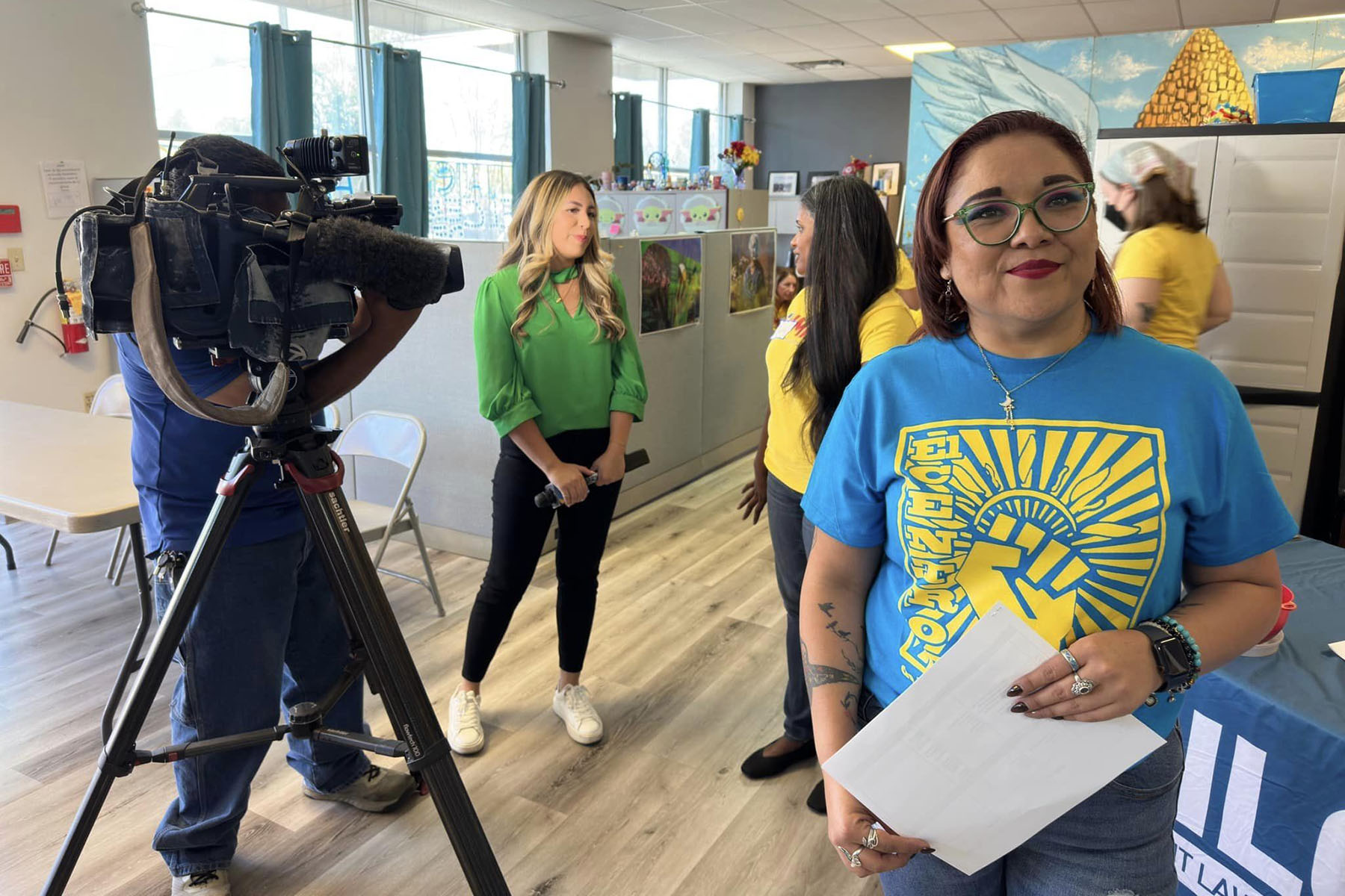 Fabiola Landeros sonríe mientras una cámara de noticias filma una entrevista detrás de ella.  Lleva una camisa azul con un puño levantado que dice "El Centro."