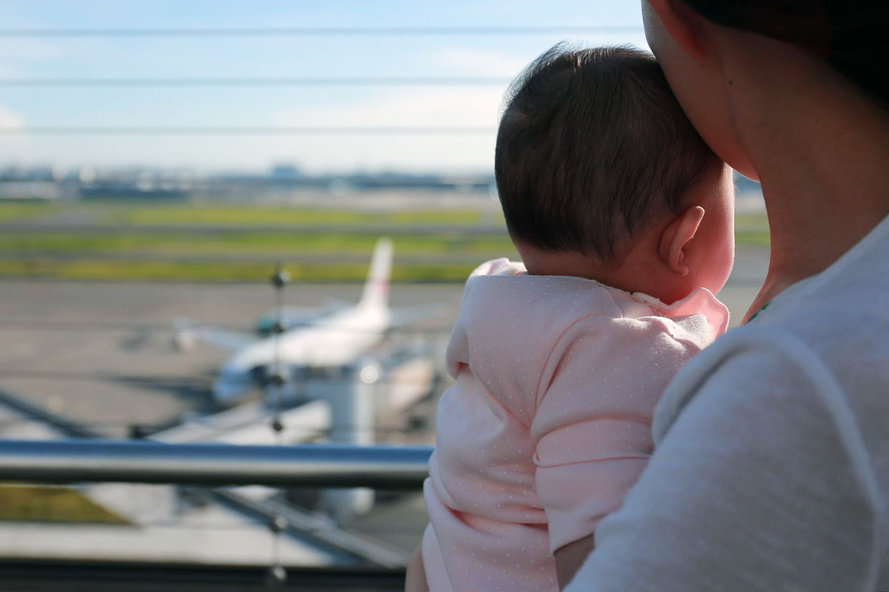 A mother holds her baby at the airport. In the background, the tarmac is seen through a window.