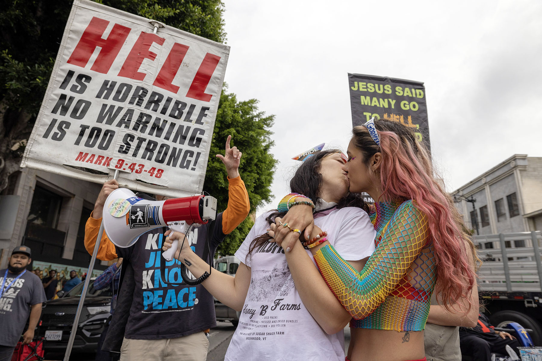 Two women kiss in reaction to confrontational Evangelical Christians condemning the annual LA Pride Parade. Behind them, a protester holds a large panel that reads "Hell is horrible, no warning is too strong!"