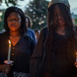 Two Black women close their eyes while holding candles during a candlelight vigil for Sonya Massey.