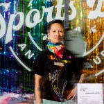 A portrait of Jenny Nguyen in front of her bar, The Sports Bra. The window is decorated with rainbow colored tassels.