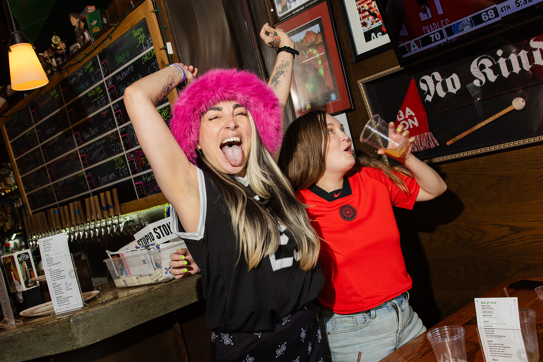 Two patrons celebrate as they watch a game inside the bar. One sticks their tongue out and one drinks a beer as they cheer.