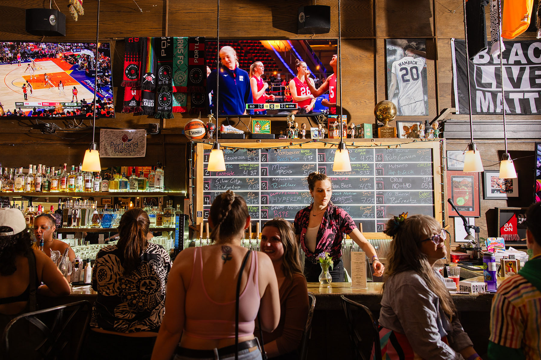 A WNBA all star game plays as patrons sit and chat at the bar. A Black Lives Matter flag and sports memorabilia hangs behind the bar.