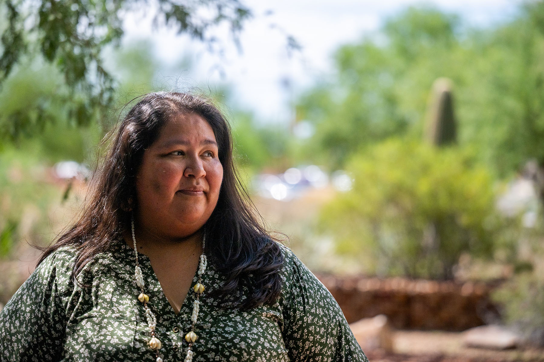 Gabriella Cázares-Kelly, wearing a green patterned blouse and a beaded necklace, stands outdoors. She looks off to the side with a thoughtful expression. Trees and greenery are visible in the blurred background.