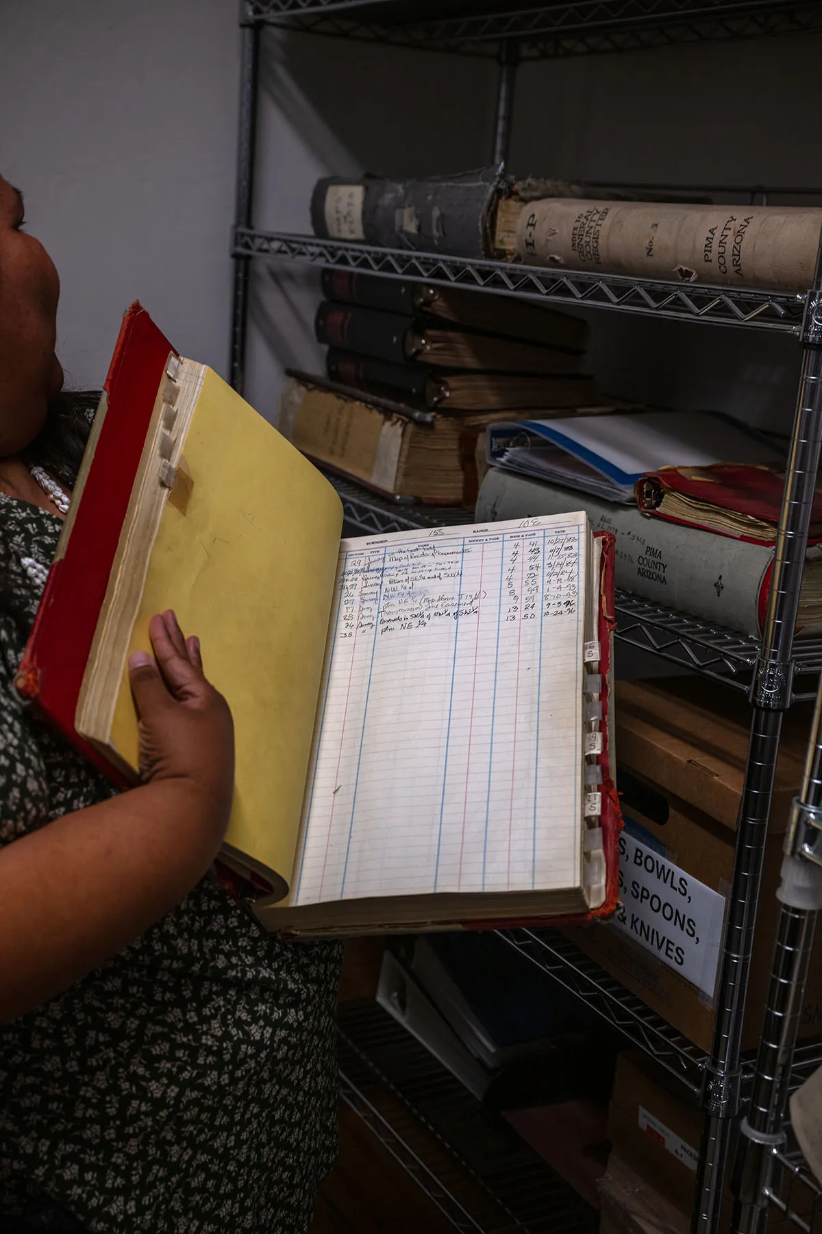 Gabriella Cázares-Kelly, partially visible, holds an open, large ledger book filled with handwritten entries. She stands in front of shelves filled with old, worn books and binders in a storage room.