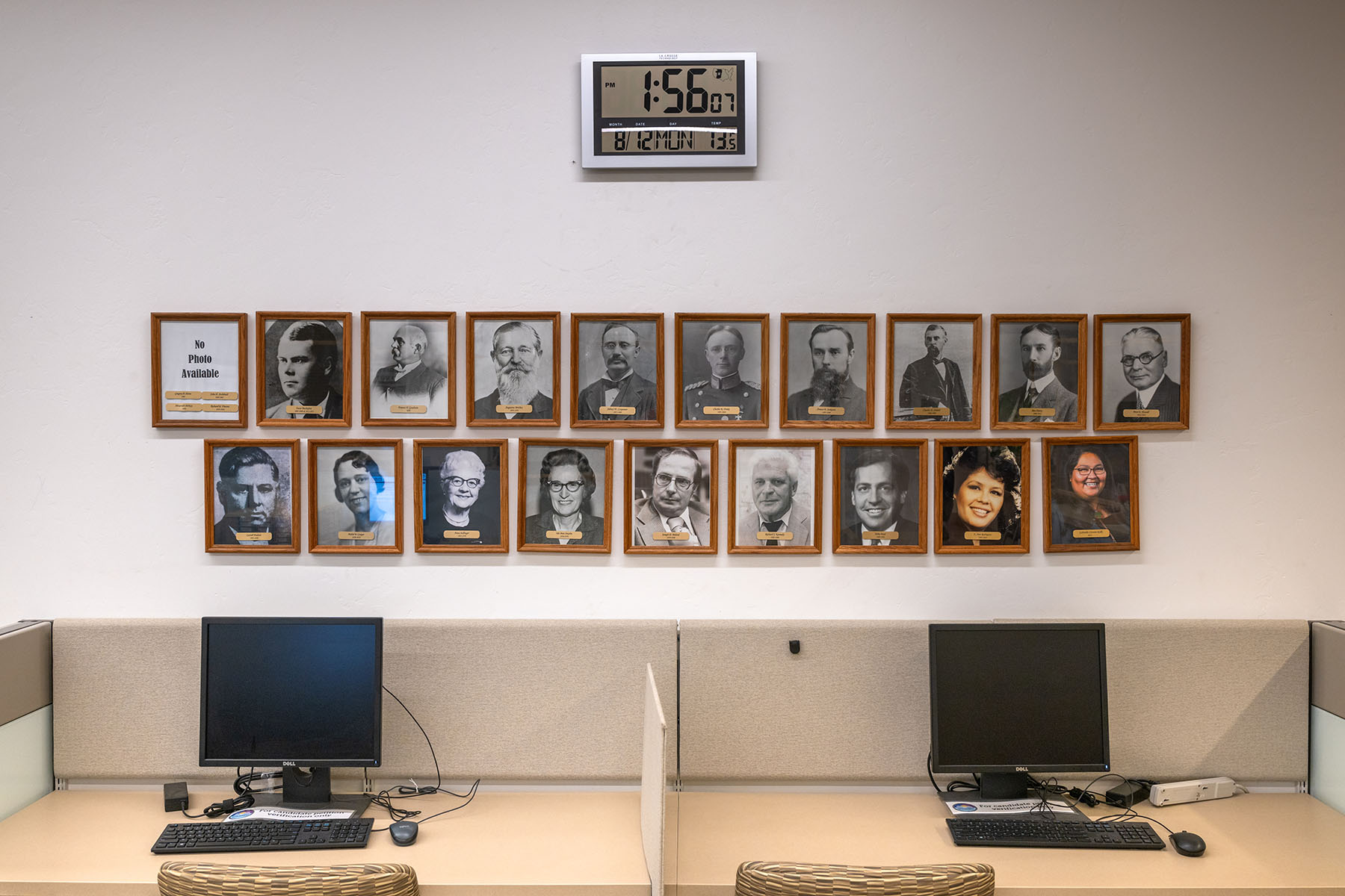 A wall in an office space displays framed portraits of county recorders, including Gabriella Cázares-Kelly, whose photo is in the bottom row. A digital clock above the frames shows the current time.