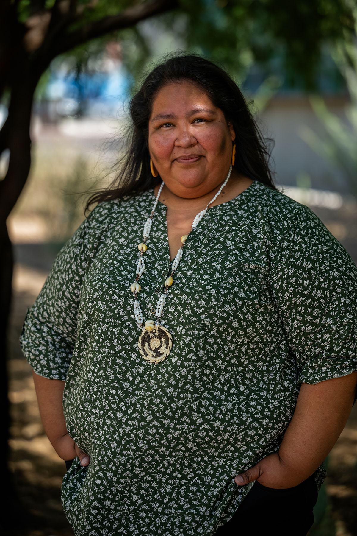 Gabriella Cázares-Kelly stands outdoors, wearing a green patterned blouse and a beaded necklace with her hands in her pockets. She smiles slightly, and trees create dappled light on her face and clothing.