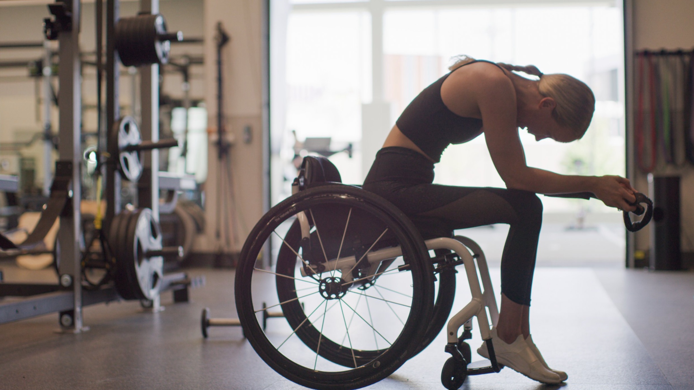 Mallory Weggemann lleva ropa deportiva y está sentada en una silla de ruedas en un gimnasio, con la cabeza agachada y las manos entrelazadas en un momento de reflexión. Está rodeada de equipos de gimnasia, incluidos bancos y estanterías de pesas, con la luz entrando a raudales por una ventana en el fondo.