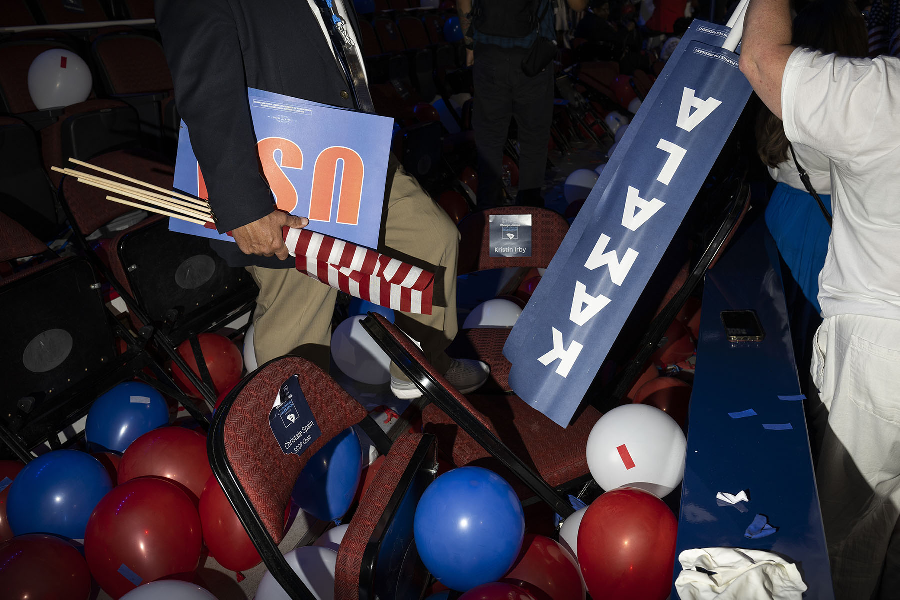 A person stands among a mess of overturned chairs and scattered red, white, and blue balloons, holding a folded blue sign that reads "USA" and a partially visible "KAMALA" sign at the conclusion of the DNC.