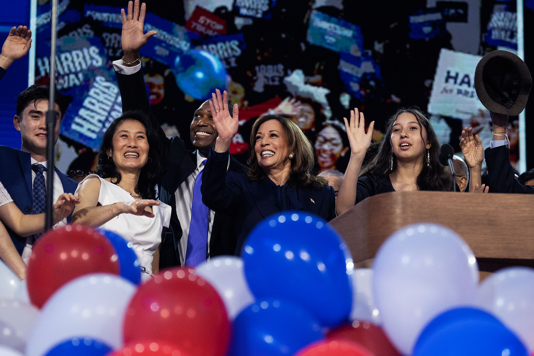 Vice President Kamala Harris is seen smiling and waving from a stage, surrounded by her family and extended family who are also waving and cheering.