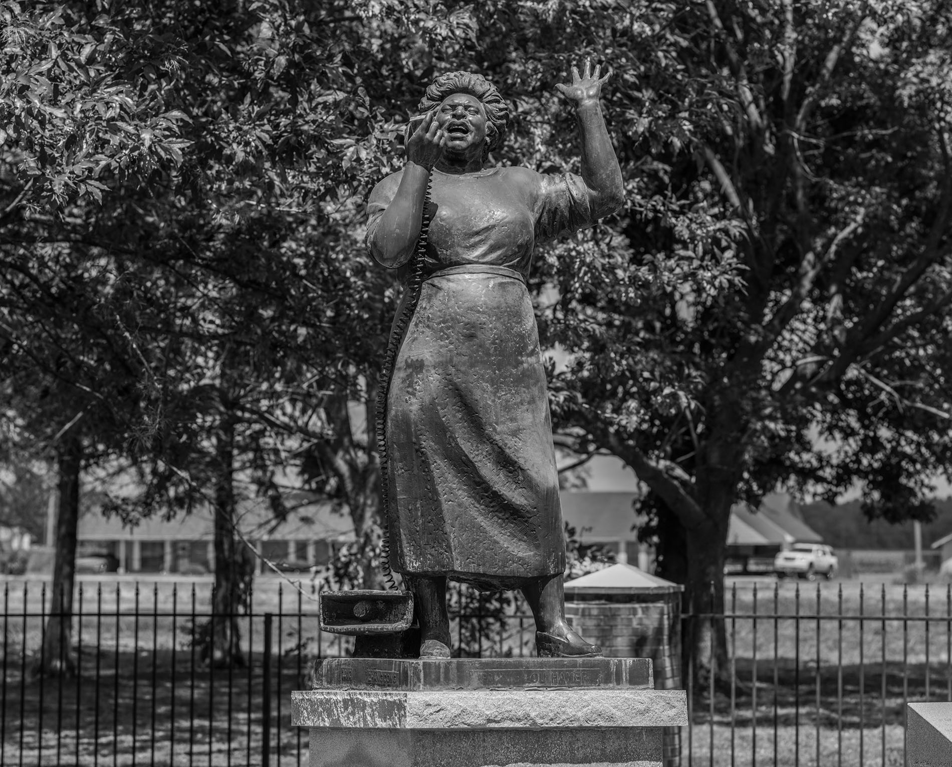 A black-and-white photograph of a statue depicting Fannie Lou Hamer with her left hand raised and her right hand near her mouth, in the midst of an impassioned speech. The statue is set outdoors, surrounded by trees, with a fenced area and a building visible in the background.