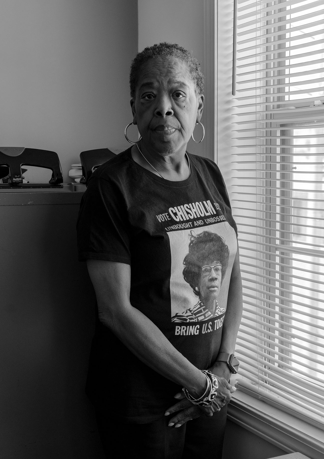 A black-and-white photograph of an older woman standing indoors near a window. She is wearing a t-shirt featuring an image of Shirley Chisholm and the text "Vote Chisholm '72 - Unbought and Unbossed." The woman is looking directly at the camera, with her hands folded in front of her.