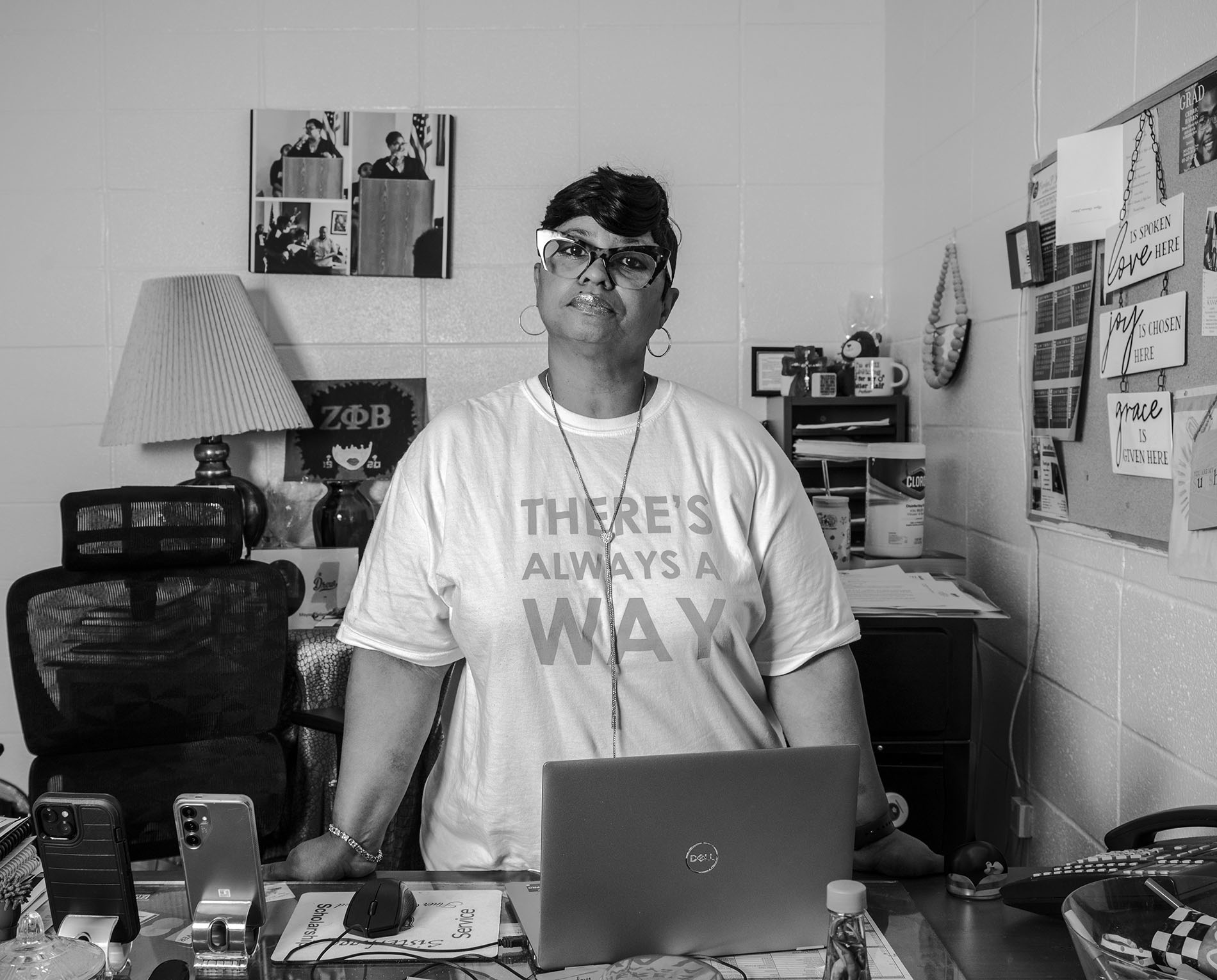 A black-and-white photograph of a woman standing behind a desk in an office. She is wearing glasses, a headband, and a t-shirt that reads "There's always a way." The desk is cluttered with papers, electronic devices, and personal items. A bulletin board and a lamp are visible in the background.