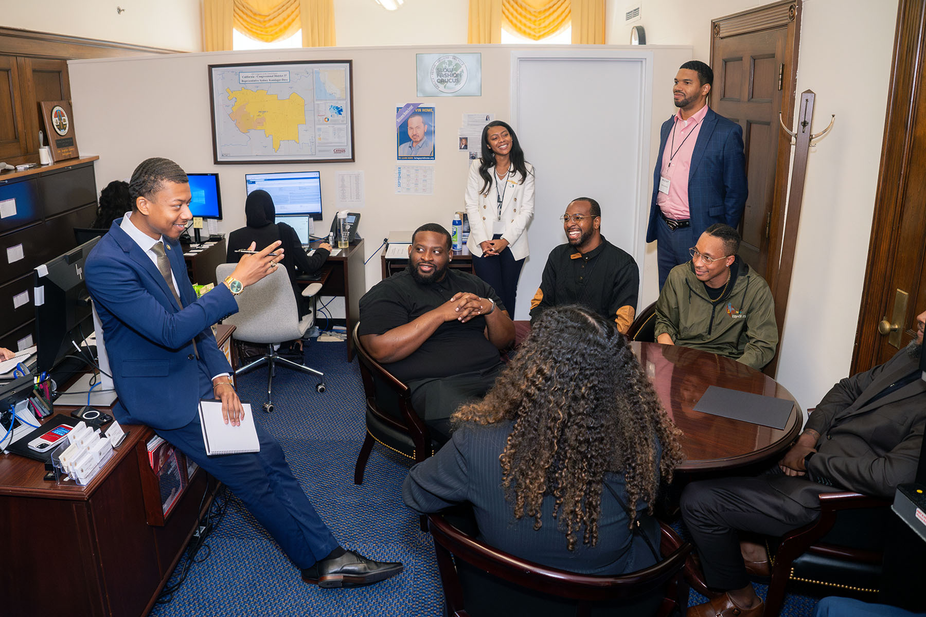 Several people sit and stand together, talking around a table in an office.