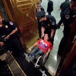 Colleen Flanagan wears a red shirt as she is being forcibly removed from the Capitol building by a police officer who is holding the back of her wheelchair. She appears to be shouting. Several officers are nearby, one of them holding a camera.