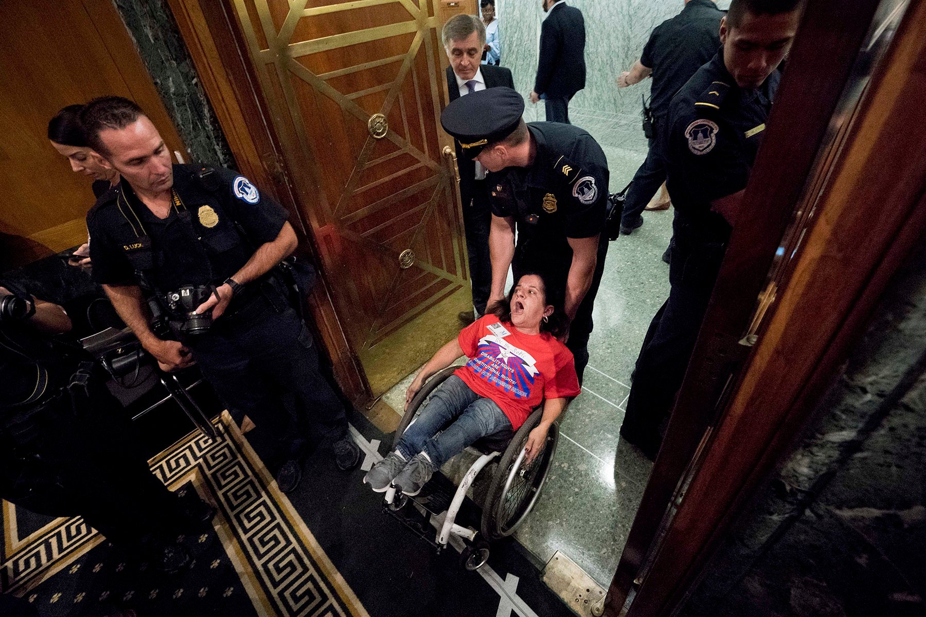 Colleen Flanagan wears a red shirt as she is being forcibly removed from the Capitol building by a police officer who is holding the back of her wheelchair. She appears to be shouting. Several officers are nearby, one of them holding a camera.