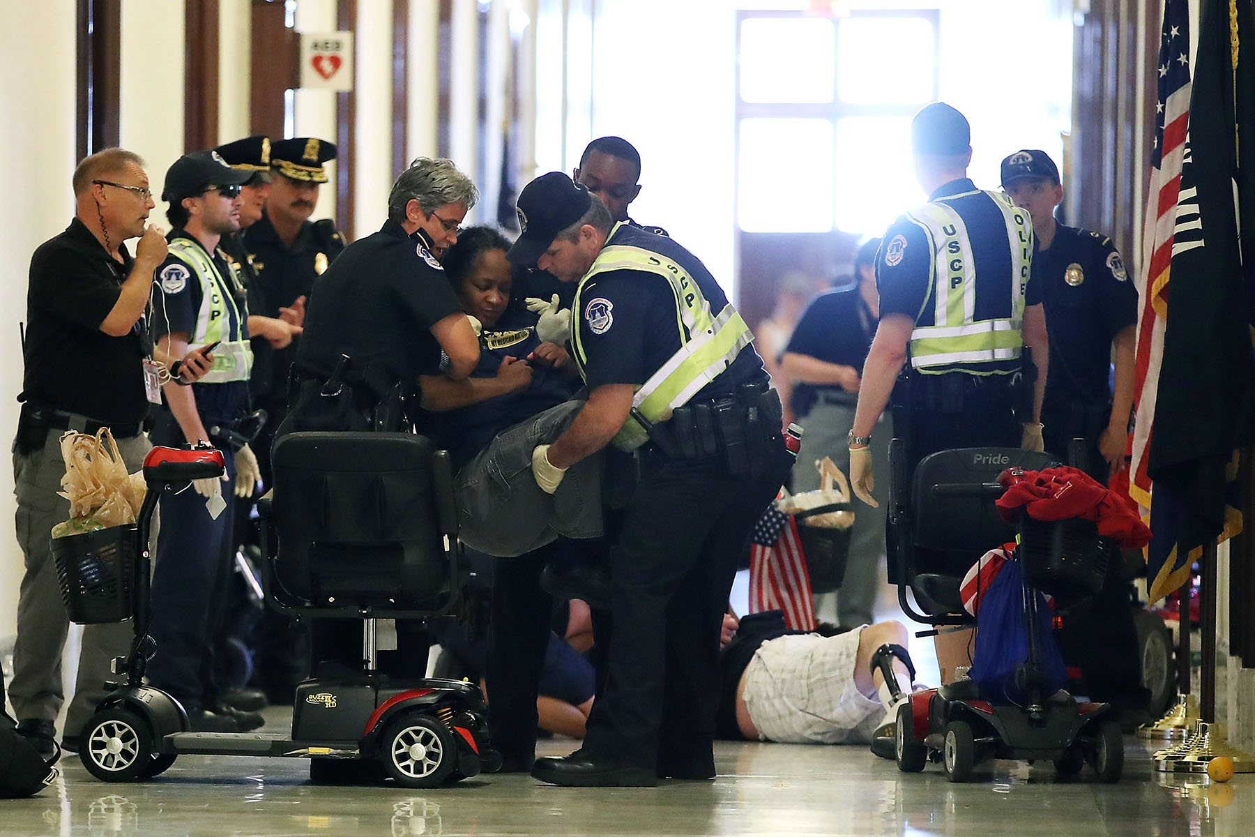 A group of police officers is forcibly removing protesters from a hallway. Anita Cameron is being lifted by two officers, while other officers stand nearby or attend to other protesters. In the background, a man lies on the floor, and mobility scooters and personal belongings are scattered around.