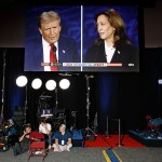 A large screen displaying former President Donald Trump on the left and Vice President Kamala Harris on the right, during the 2024 presidential debate. The screen shows a live broadcast by ABC News. Below the screen, three crew members are seated on the floor with various camera and lighting equipment around them in the media center at the Pennsylvania Convention Center.