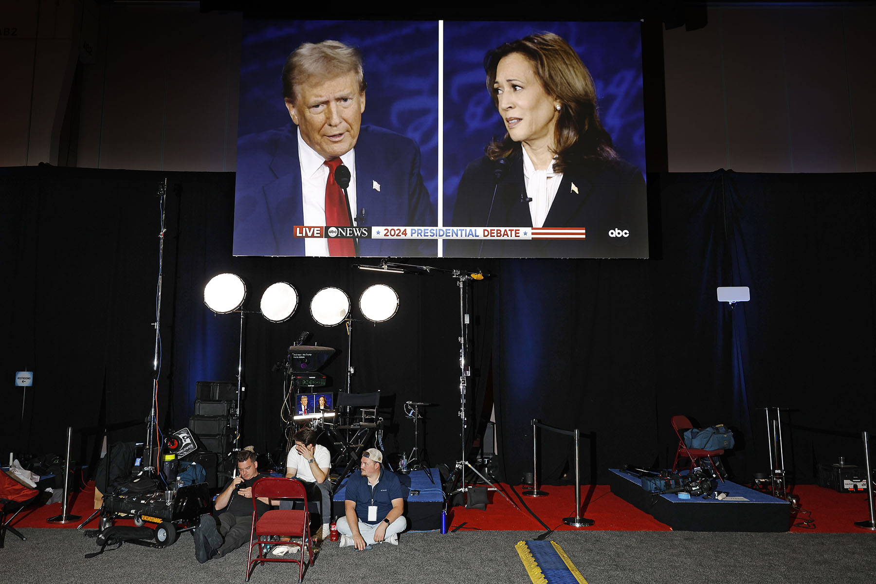 A large screen displaying former President Donald Trump on the left and Vice President Kamala Harris on the right, during the 2024 presidential debate. The screen shows a live broadcast by ABC News. Below the screen, three crew members are seated on the floor with various camera and lighting equipment around them in the media center at the Pennsylvania Convention Center.