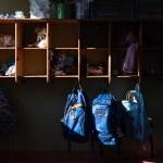 A set of cubbies and hooks are seen at a day care center. Each cubby contains various items like hats, scarves, and toys, while several backpacks hang from hooks below.