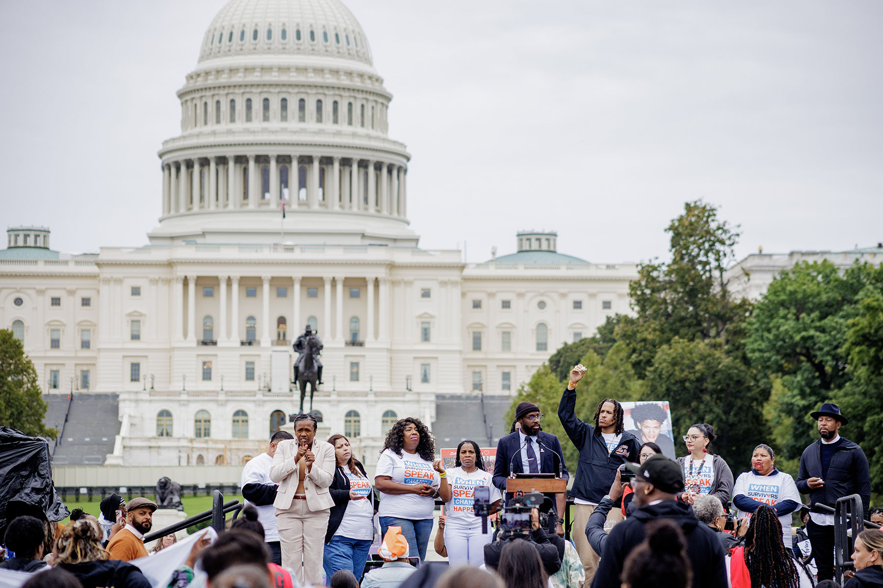 Un grupo de oradores se encuentra frente al Capitolio de los Estados Unidos durante la Marcha del Habla de Sobrevivientes del Crimen en Washington, DC. Los oradores están rodeados por una gran multitud, y una persona levanta el puño en el aire. El edificio del Capitolio se ve al fondo.