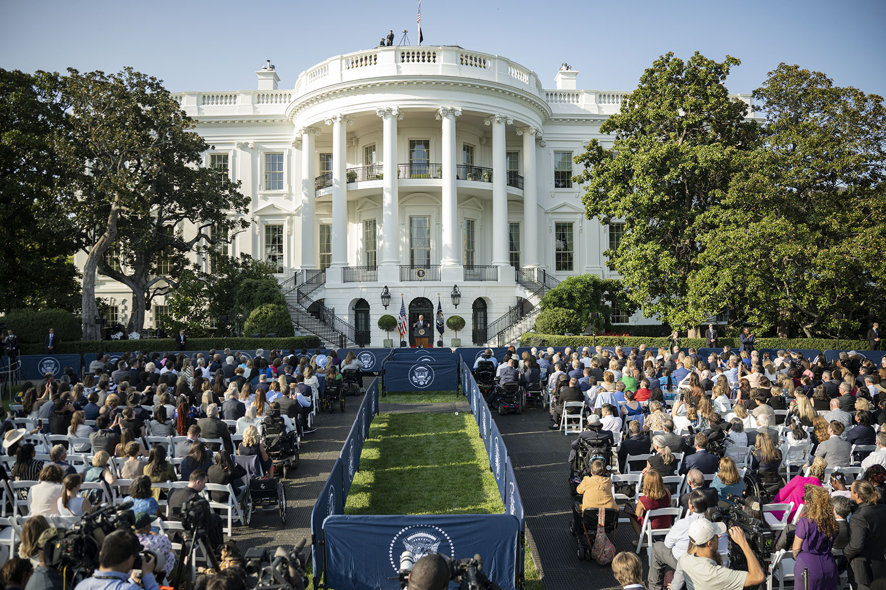 President Joe Biden delivers remarks to celebrate the Americans with Disabilities Act and to mark Disability Pride Month on the South Lawn of the White House. In the foreground, a crowd of diverse attendees, some in wheelchairs, listens to him speak at a podium positioned in the center.