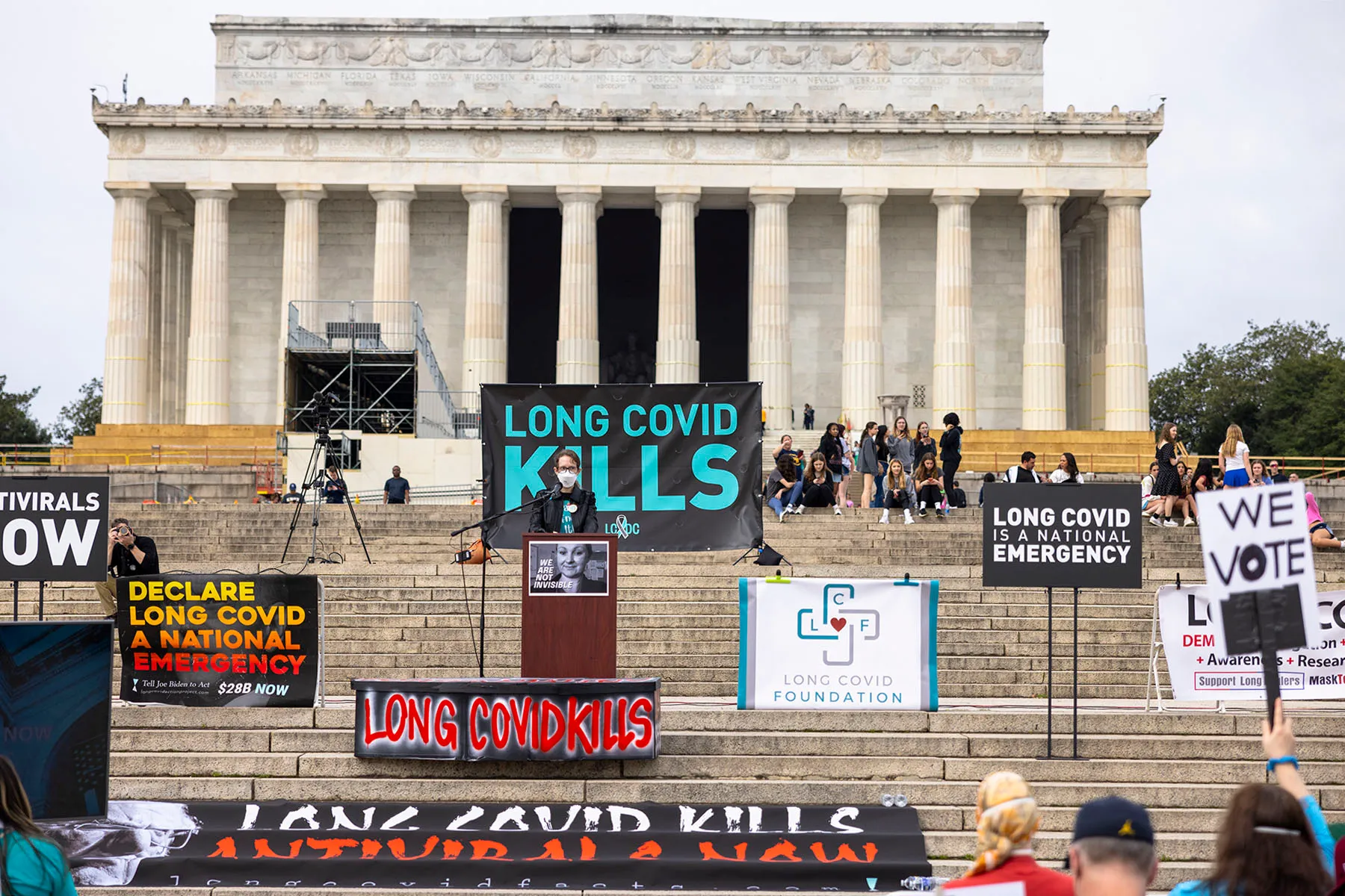 Renee Addams, a Long COVID advocate, speaks at a podium in front of the Lincoln Memorial in Washington, D.C. Signs around her read "Long COVID Kills" and "Declare Long COVID a National Emergency."