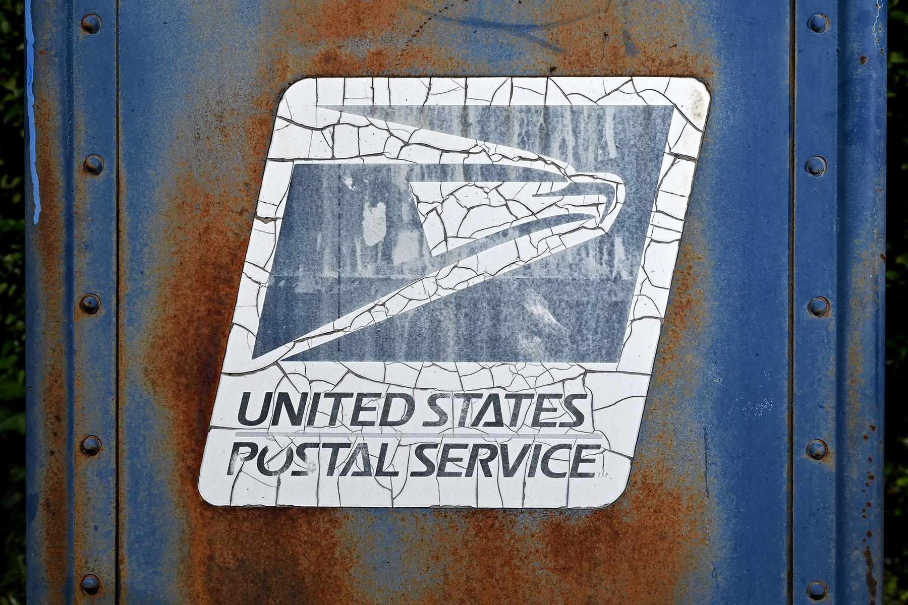 A close-up of a weathered and rusty United States Postal Service (USPS) mailbox. The USPS logo, featuring an eagle, is visibly cracked and peeling on the surface.