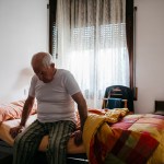 An elderly man sits at the edge of a bed alone in a room with a window at his back.