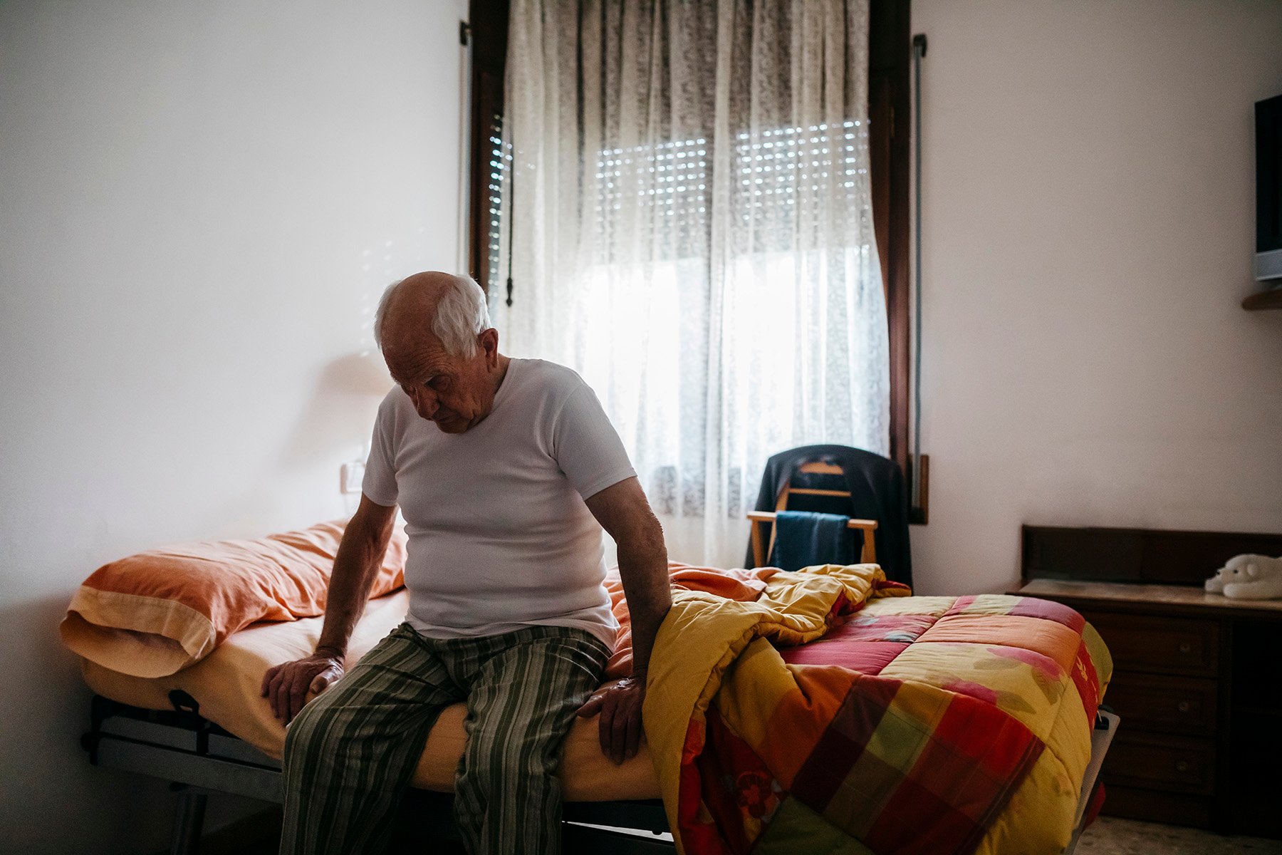 An elderly man sits at the edge of a bed alone in a room with a window at his back.