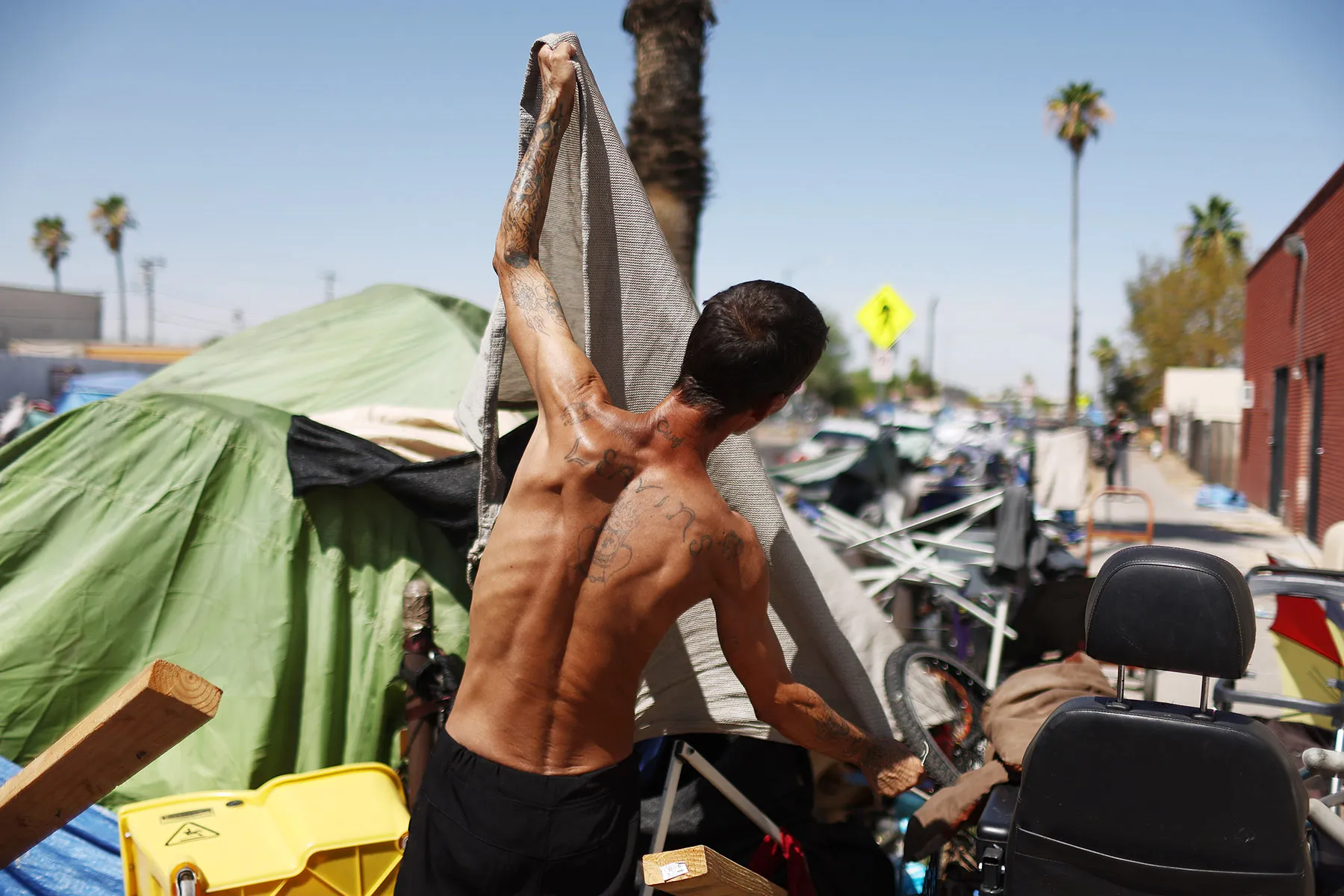 A shirtless person faces away from the camera as they stretch out a tarp near a tent set up on a street.