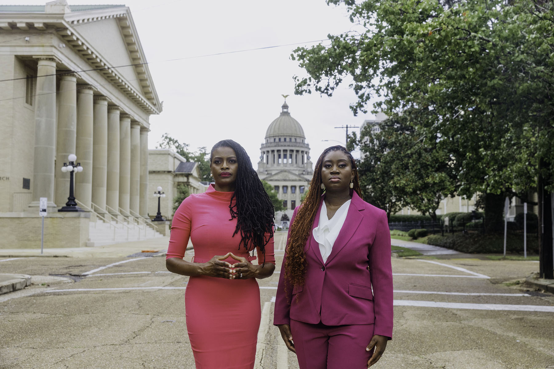 Cassandra Welchlin and Velvet Scott standing side by side in front of the Mississippi Capitol building. Cassandra, on the left, is wearing a bright pink dress, and Velvet, on the right, is dressed in a pink suit.