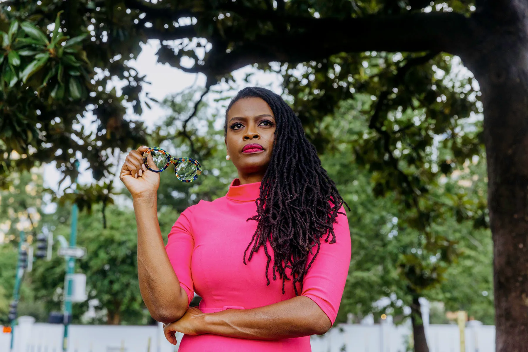 Cassandra Welchlin holding glasses in one hand, standing under a tree with a determined expression. She is wearing a bright pink dress, with her long locs draped over her shoulder, and the background features a park setting.