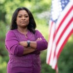 Diamond Staton-Williams, wearing a purple sweater, stands with arms crossed in front of an American flag.