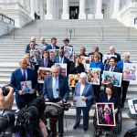 Sen. Chuck Schumer, flanked by Sens. Cory Booker, Patty Murray and Tammy Duckworth, speaks into a microphone on the Senate steps with other Senate Democrats behind him, many holding large-format photos.