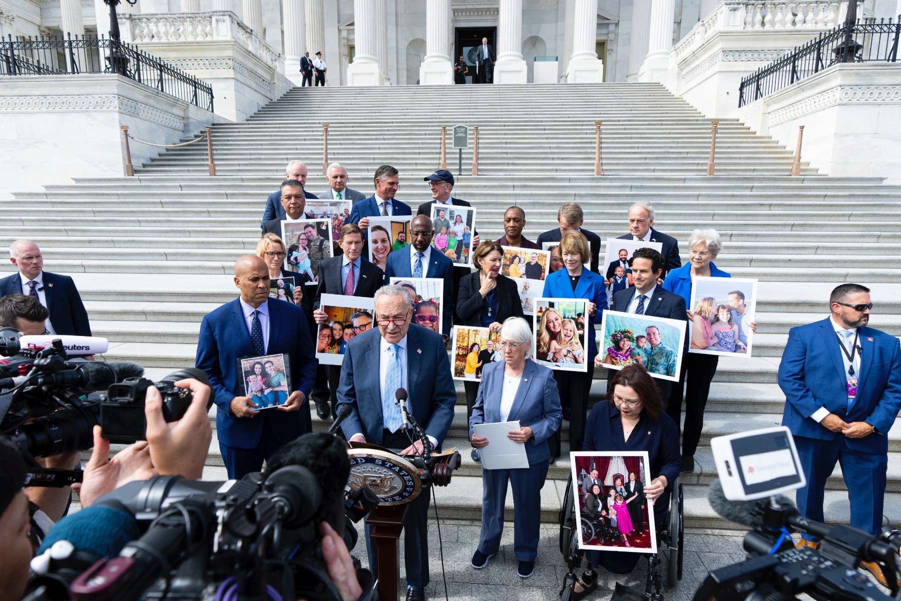 Sen. Chuck Schumer, flanked by Sens. Cory Booker, Patty Murray and Tammy Duckworth, speaks into a microphone on the Senate steps with other Senate Democrats behind him, many holding large-format photos.